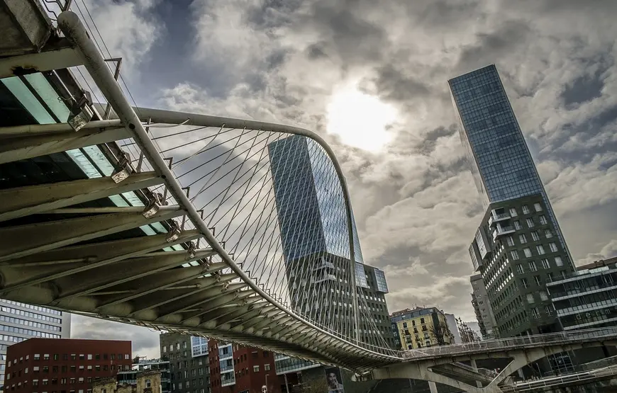 un-ponte-bianco-con-una-forma-contemporanea-disegna-delle-linee-curviformi-di-fronte-alle-due-torri-di-bilbao